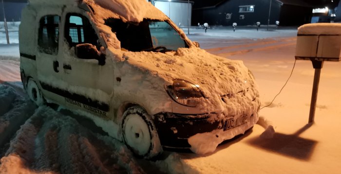 Skåpbil täckt med snö, parkerad nattetid, elektrisk motorvärmare inkopplad, kallt väderförhållande, belysning från gatulampor.