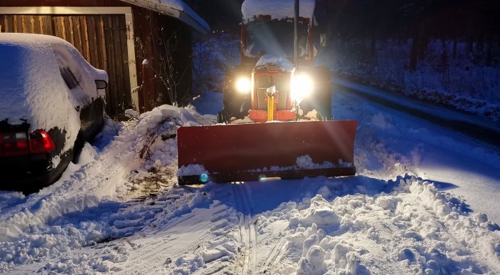 Traktor med snöblad plogar snö nära parkerad bil, kvällstid, vinterlandskap, upplyst av strålkastare.