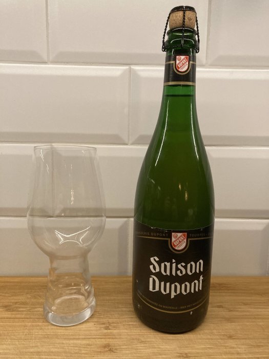 En grön flaska Saison Dupont-öl med kork och muselet, bredvid ett tomt ölglas på träbänk.