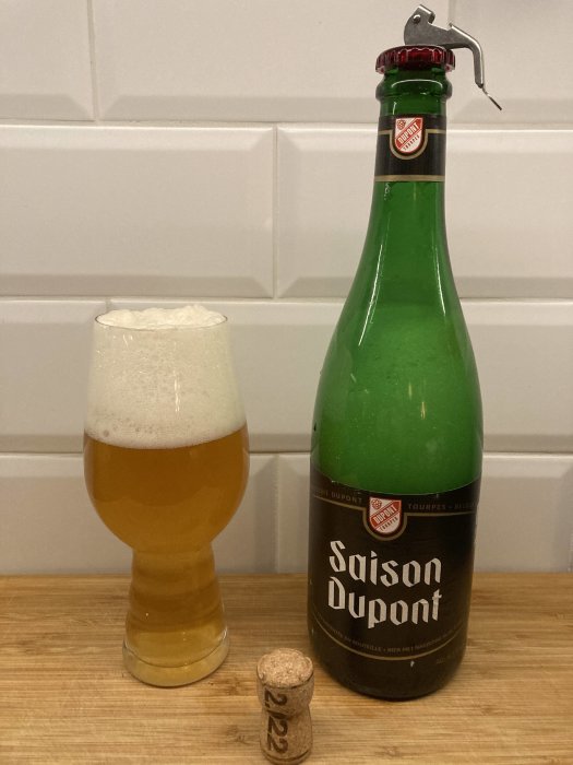 Öppnad Saison Dupont ölflaska med glas och kork på träbänk, kakelbakgrund.