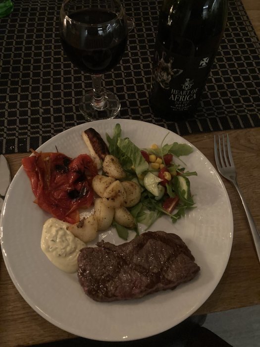 Middagstallrik med biff, grönsaker, grillade tomater, potatis, sås och ett glas rödvin samt en vinflaska.