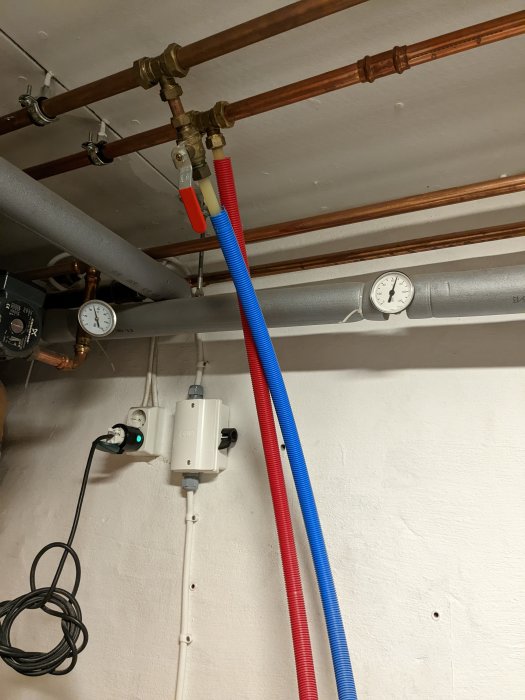 VVS-installation med kopparledningar, röda och blå slangar, manometer, eluttag och kablar på en vägg.