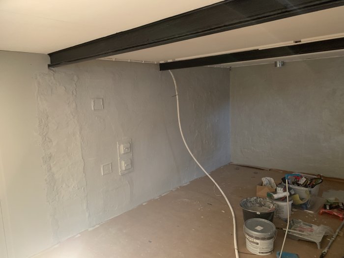 Ett rum under renovering med oslipade väggar, kablar, färgburkar och byggmaterial på golvet.