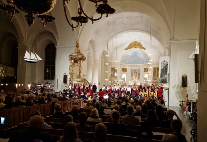 Kör och orkester framför musik för publik i en ljus kyrksal, inredd med ljuskronor och pendat.