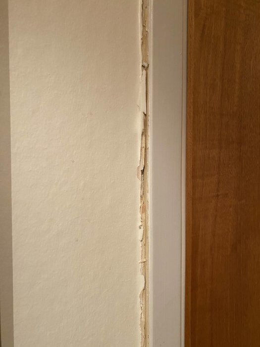Skadad väggkant vid dörröppning, flagnande färg, behov av reparation, trä och vit ytbehandling.