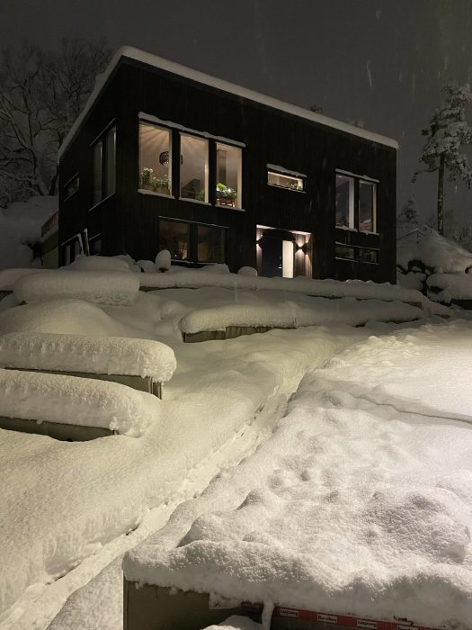 Modernt hus med tända fönster, omgivet av djup snö om natten. Träd syns i bakgrunden.