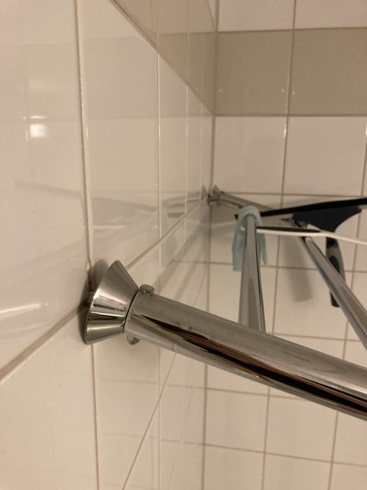 Metallisk handdukshängare, vita kakelväggar, hörna på badrumsvägg, del av duschstång synlig.