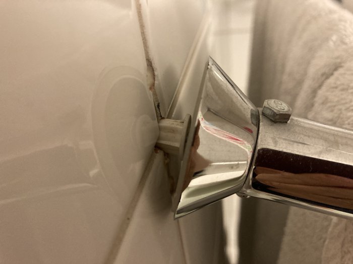 Duschväggskant möter vita kakelväggen, synlig smuts, skruv och duschväggsfäste i metall och glas.