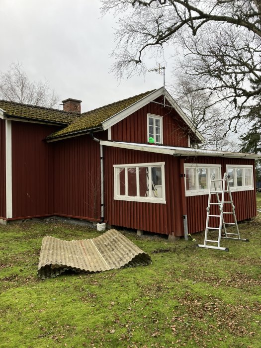 Rött trähus med vita knutar, eternittak, stege, lövträd, gräsmatta, liggande eternitplatta.