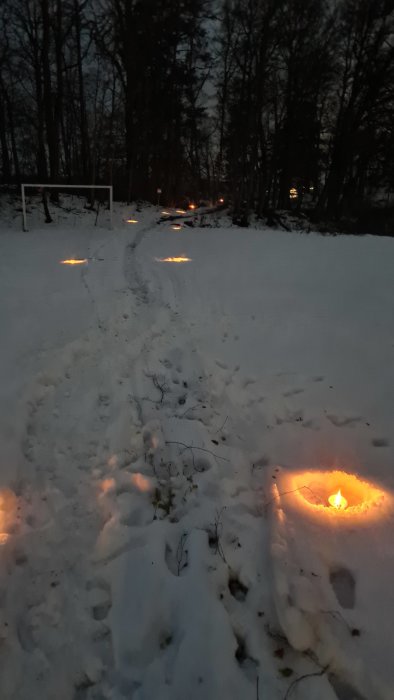 Ett snöigt landskap i skymningen med ljus längs en stig och fotspår i snön.