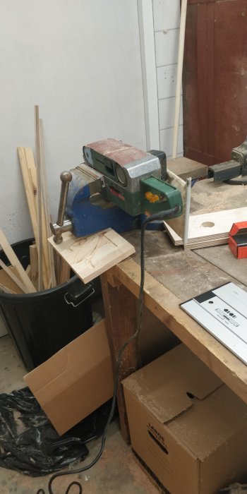 Verkstadsbord med bänkslipmaskin, träbitar, lådor, och snickeriverktyg; oorganiserat med sågspån.