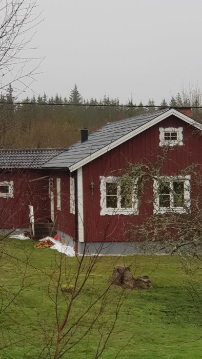 Röd stuga med vita knutar, skog i bakgrunden, molnigt, tidig vår, grönt gräs, utan solsken.