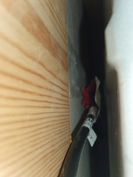 Kabel och träyta i smalt mellanrum. Svårdefinierbar röd föremål syns i djupet. Otydlig, mörk omgivning.