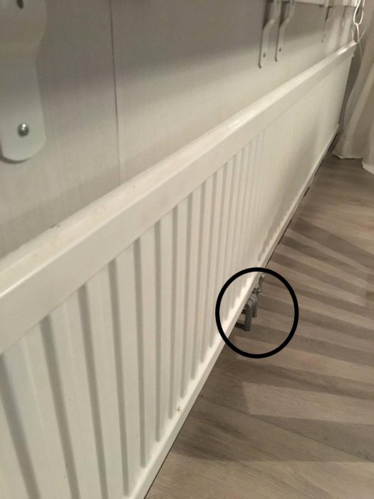 Element i ett rum, vit väggmonterad radiator, trägolv, nedre delen av bilden markerad med cirkel.