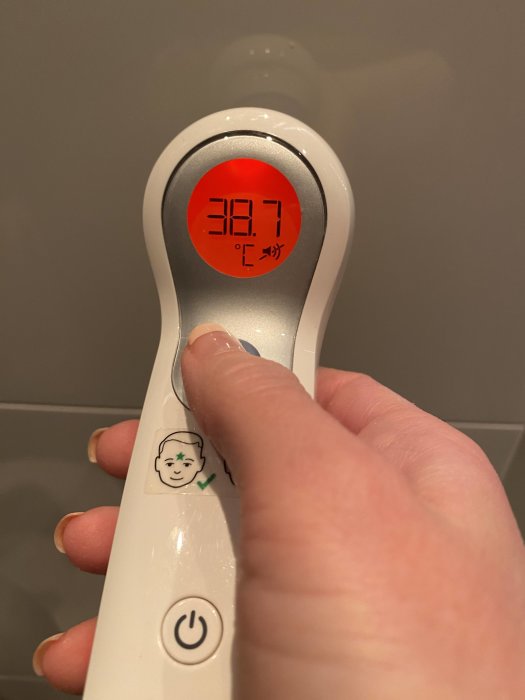 Infraröd termometer visar feber temperatur på 38.7 grader Celsius hållen av en hand.