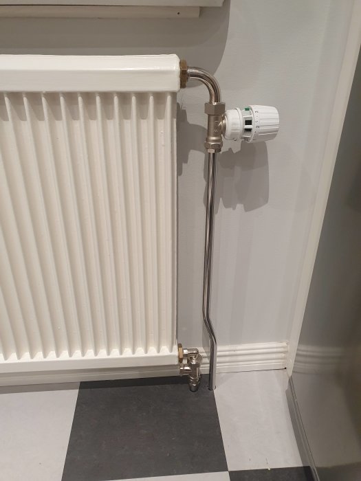 Vit radiator med termostat, böjda rör, svartvit rutigt golv, en vit vägg.