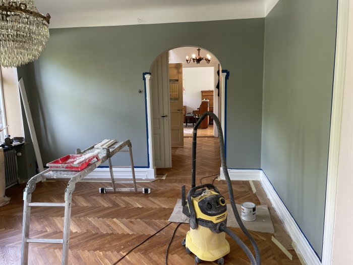 Renoveringsarbete i rum, målarverktyg och tejp, dammsugare, stege, öppen dörr till annat rum.