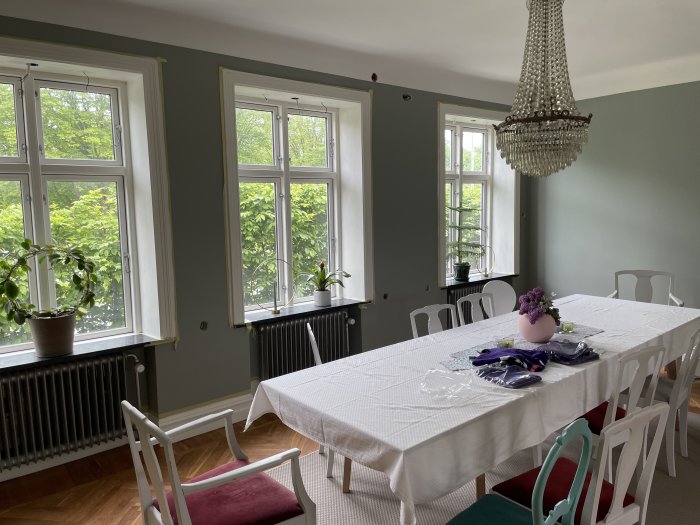 Ett matsalsrum med vitt bord, stolar, kristallkrona och fönster med utsikt mot grönska.