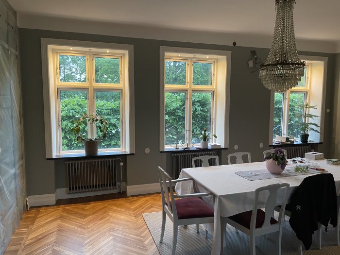 Matsal med trägolv, bord, stolar, kristallkrona, fönster med utsikt över grönska, vit gardin, och radiatorer.