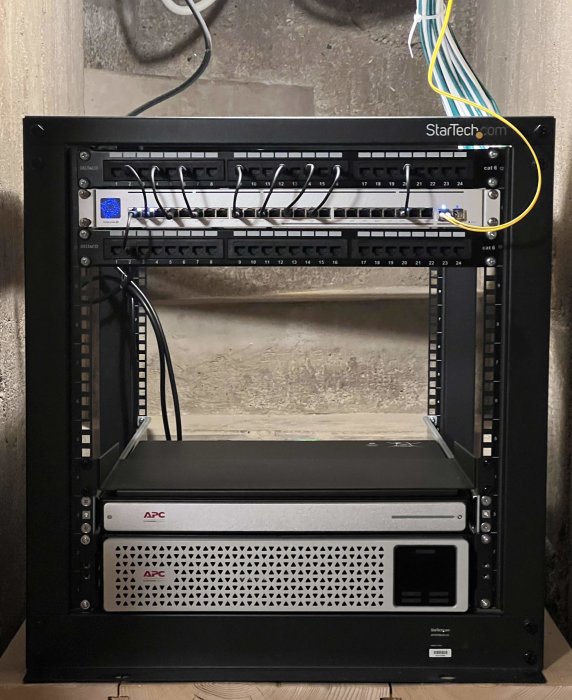 Serverrack med nätverksutrustning, patchpaneler, och UPS från APC, monterat mot betongvägg.