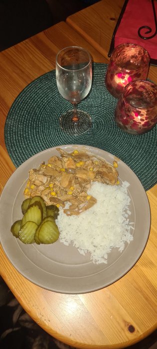 Middagsdukning med ris, stroganoff, saltgurka, dricksglas och ljuslyktor på ett träbord. Hemtrevlig atmosfär.