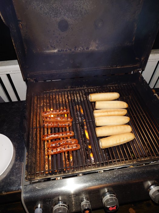 En grill med korvar och bröd, flammor, grillverktyg, kvällstid på veranda.