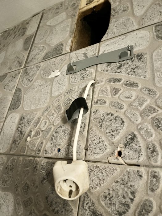 Vägg med kakel, trasig eluttag, tejp, osäker elektrisk säkerhet, renoveringsbehov, sladdar, hål i vägg.