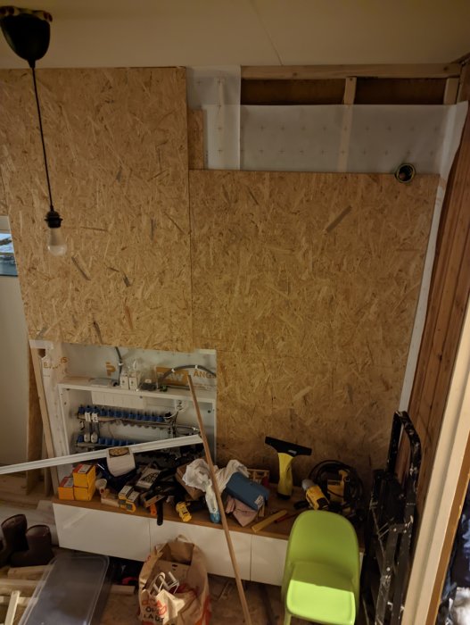 Oorganiserad arbetsplats, verktyg, byggmaterial, OSB-vägg, renovering pågår, rörledningar synliga, oavslutat utrymme.