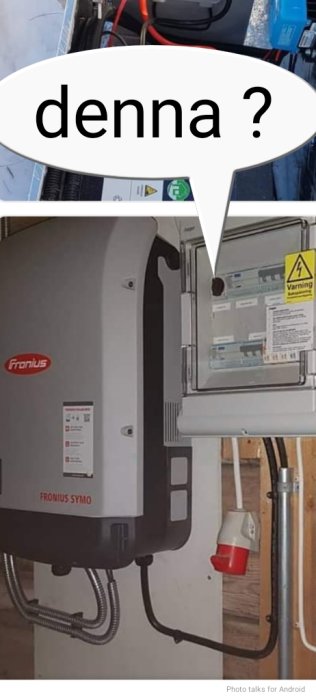 Fronius Symo solenergi-inverter, installationspanel, elektriska kopplingar, rör, varningsetiketter, ser ut som ett garage eller teknikrum.