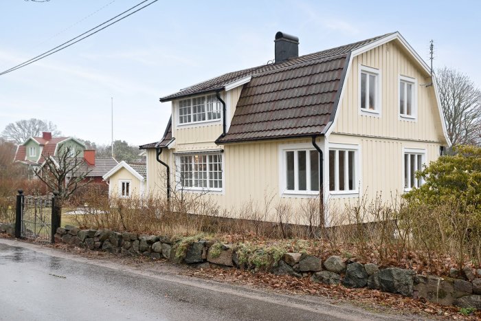 Gult trähus med tegeltak, stenmur, grå himmel, fuktig dag, landsväg framför.