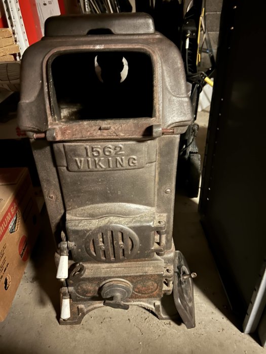 Gammal, dammig vedspis märkt "1562 VIKING", står i ett rörigt förrådsutrymme med olika föremål.