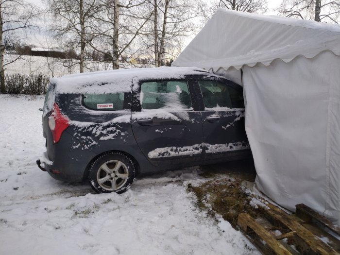 Svart bil parkerad under vit tältgarage i snölanskap, delvis täckt av snö, omgiven av träd.