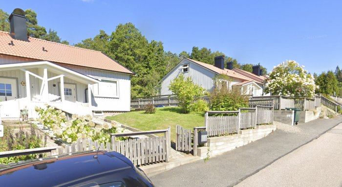 Soligt bostadsområde med enfamiljshus, grönska, staket, bil och klarblå himmel.