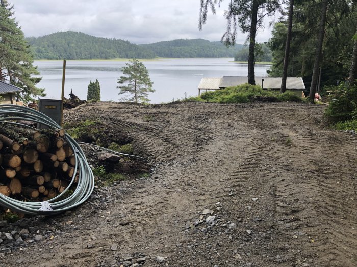 Tomtmark nära vatten med grustäckt yta, timmerhög och kabelrulle, skog och molnig himmel i bakgrunden.