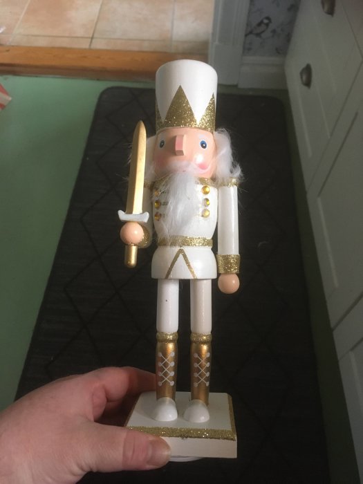 En hand håller en julnötknäppare i vitt och guld, traditionell prydnadsfigur, står på en köksgolv.