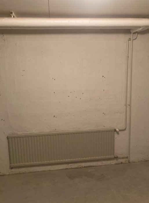 Källarlokal med vitmålad vägg, radiator, rörinstallation och synligt slitage.