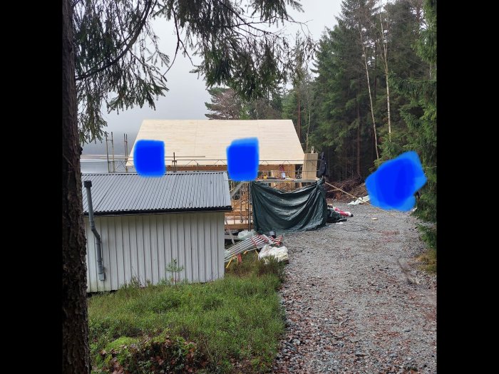 Byggarbetsplats i skogsmiljö, halvfärdigt trähus, skyddsdukar, grusväg och ett skjul. (Blåa fläckar täcker delar av bilden.)