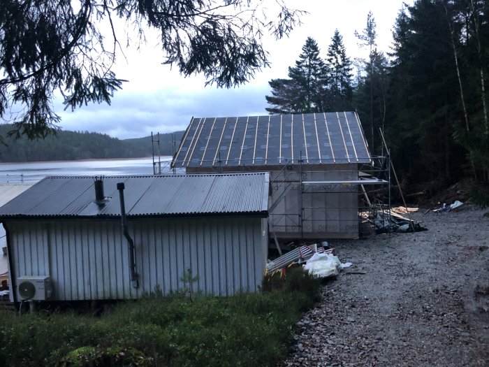 Under konstruktion, hus med solpaneler, skog, sjö, gråmulen himmel, stillsam naturmiljö, byggmaterial och ställningar syns.