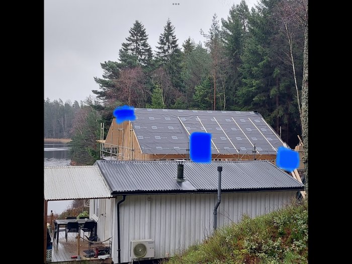 Hus under konstruktion med takduk, skog bakom, sjö i bakgrunden, grå himmel, plåttak i förgrunden.