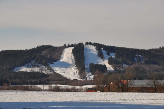 Ett snötäckt landskap med skidbackar, skog och en bondgård i förgrunden. Soligt väder.