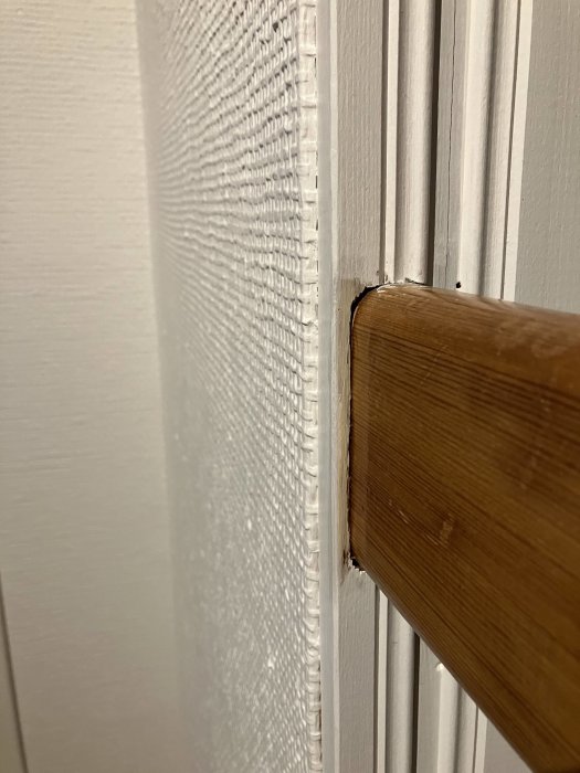 Närbild av en vit texturerad vägg, trälist, skada där list möter en dörrkarm.