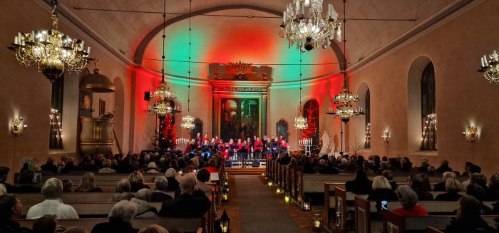 Kör uppträder i kyrka med juldekorationer, färgad belysning, och en publik lyssnar uppmärksamt.