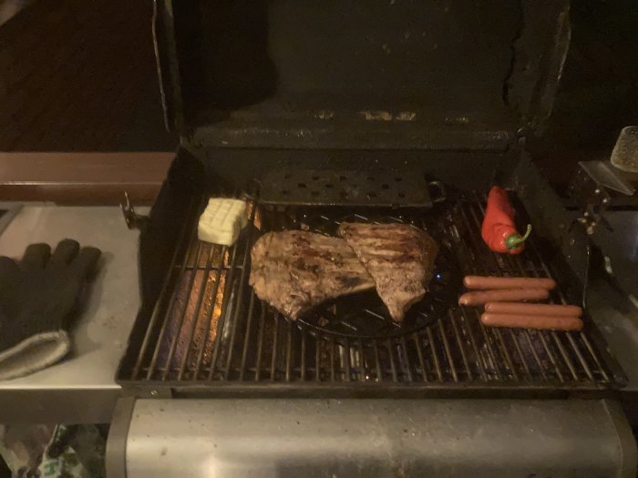 En grill med stekta köttstycken, korv, ost och en röd paprika, med grillhandske på sidan.