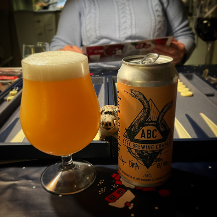Ölglas med skum, ölburk från Apex Brewing, brädspel i bakgrunden, mysig samvaro.