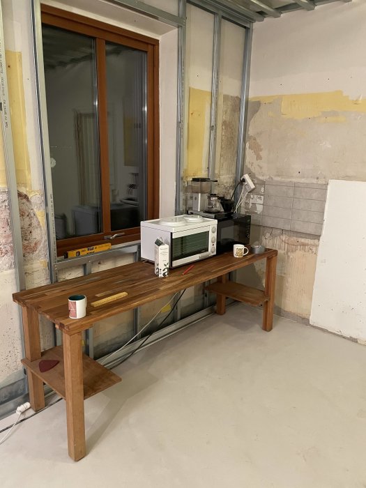 Orenoverat rum med bord, mikrovågsugn, kopp och byggmaterial. Under renovering eller ombyggnad.