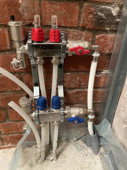 Industriellt rörverk med ventiler och mätare mot tegelvägg, installationsristningar, oavslutade byggarbeten synliga.