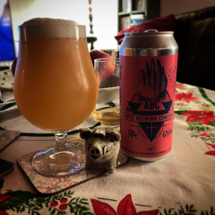 Ett grumligt öl i glas, ölburk från Apex Brewing, smågrisfigur, julduk, stämningsfull belysning.