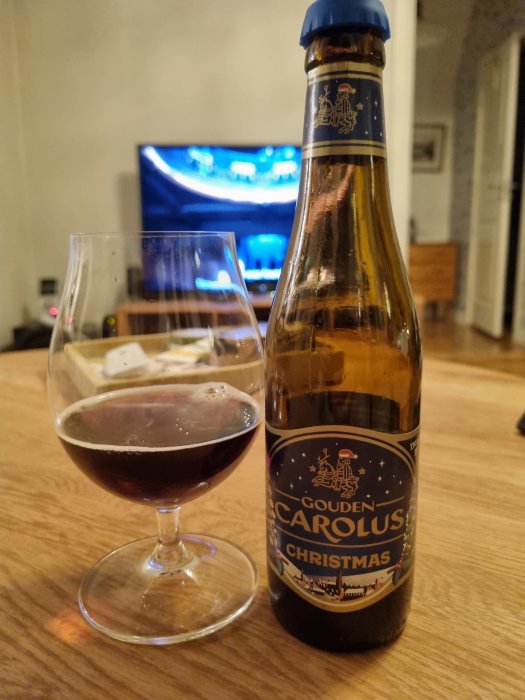 Gouden Carolus Christmas ölflaska och snifterglas, mörkt öl, suddig tv i bakgrunden, hemmiljö.