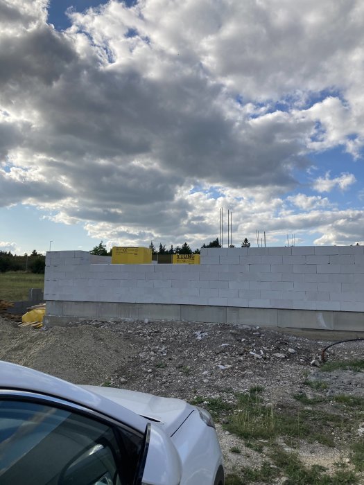 Byggarbetsplats med grundmur, betongblock, armeringsjärn, molnig himmel och bilens framsida.