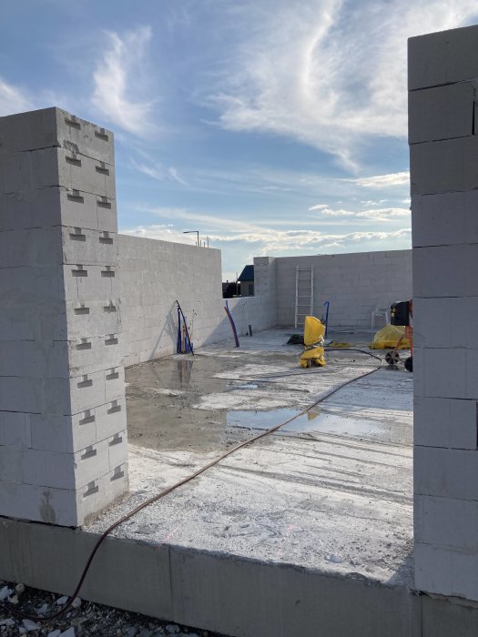 Byggarbetsplats med ofärdiga väggar av vit betongblock, blå himmel, moln, byggmaterial och stegar synliga.